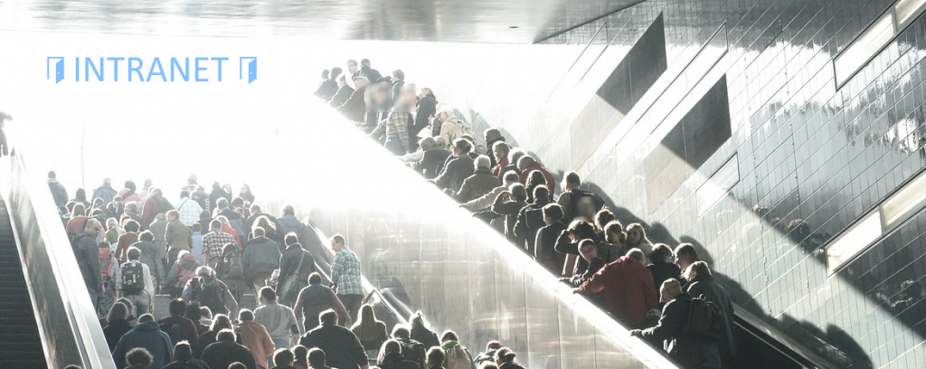 Eine Menschenmenge bewegt sich in Richtung eines Intranet-Schildes; eine Hälfte der Menschenmenge benutzt die Treppe, die andere Hälfte benutzt eine Rolltreppe.